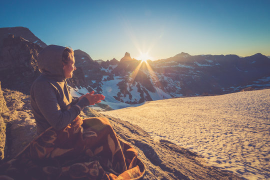 Girl sitting on a rock and covered with a blacket is watching Sunset From the top of the Mountain in a glacier area in the Alps with snow and blue sky