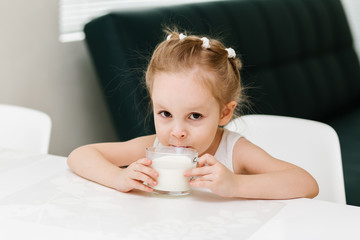 Little smart caucasian girl drinks milk from a glass cup in the kitchen and has fun, healthy eating concept for children.