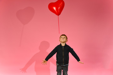 A boy throws and catches a heart-shaped balloon on a pink background in the studio.