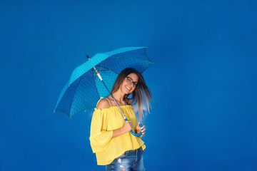 Fototapeta premium Portrait of cute teenage girl in yellow blouse holding blue umbrella with black dots while standing in front of blue background.