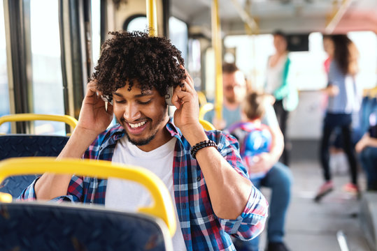 Smiling African guy sitting in the public transportation and putting on headphones. In background people sitting and standing.