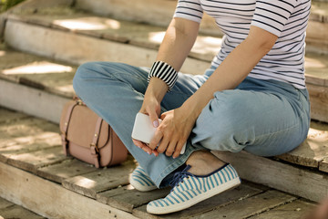 Young woman in blue jeans and striped sneakers sits on old wooden steps and holds a phone in her hands.