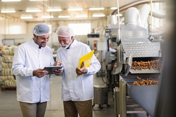 Two workers in uniforms looking at tablet while standing in food factory. Older one pointing at tablet.