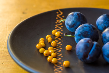 still life - juicy blue plums and sea buckthorn berries in a ceramic plate on a wooden background