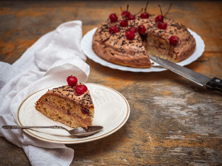 A piece of homemade cake and the cake itself in the background, decorated with red cherries, next to a white cotton napkin