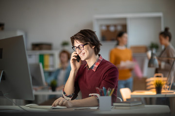 Smiling young man in casualwear sitting in front of computer monitor consulting with his colleague...