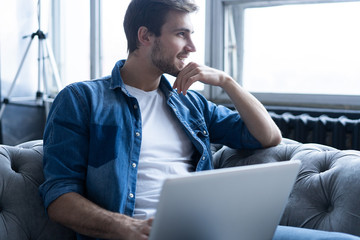 Young attractive smiling guy is browsing at his laptop, sitting at home on the gray sofa at home, wearing casual outfit.