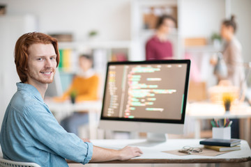 Young successful designer or programmer looking at you while sitting by desk and decoding online...