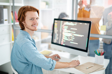 Young cheerful and successful programmer or webdesigner sitting by desk in front of computer screen...