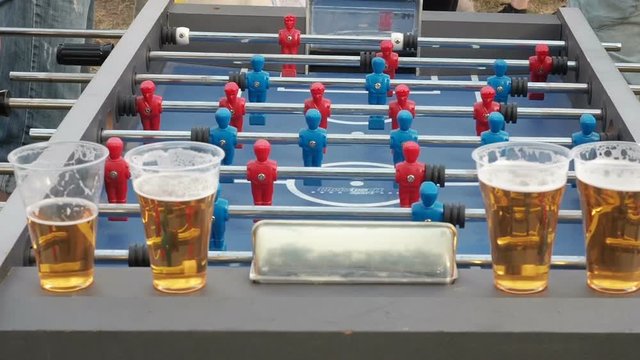 Young people play table football and drink beer outdoors. Figures of footballers move left and right people playing foosball. Table soccer plaers. Table football soccer game kicker close up. Outdoor