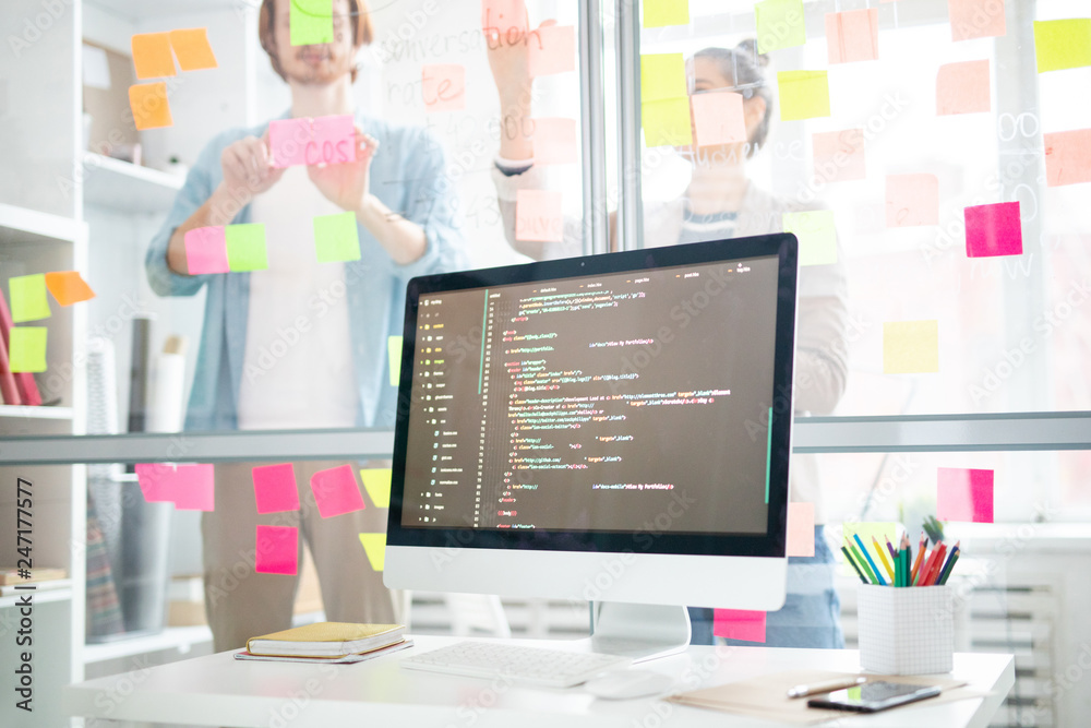 Poster Computer screen with coded data on desk on background of office workers sticking notepapers on transparent board