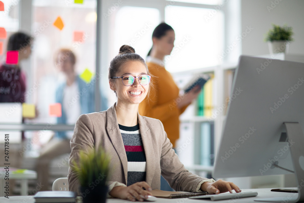 Poster happy casual businesswoman looking at you while sitting by desk in front of computer screen in worki