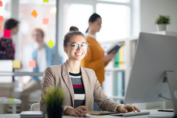 Happy casual businesswoman looking at you while sitting by desk in front of computer screen in...