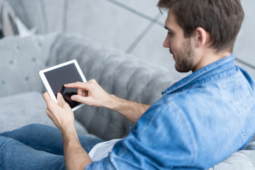 Handsome man is using a digital tablet and smiling while resting on couch at home.