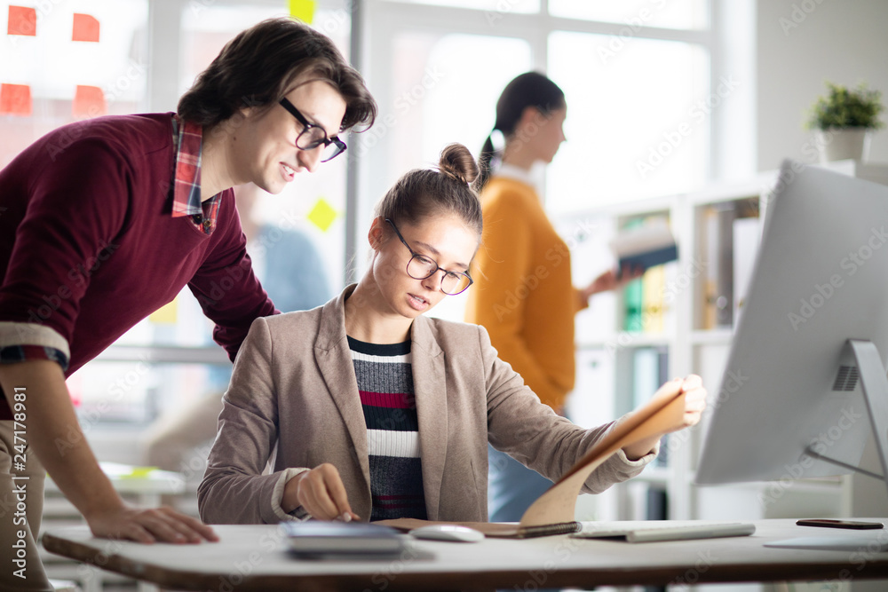Poster young businesswoman and her colleague reading working points in notepad while organizing work