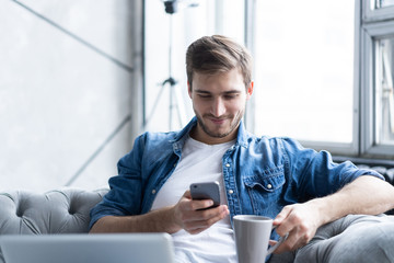 Young man using his smartphone for online banking - sitting on sofa with laptop on leap.