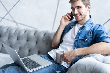 Portrait of handsome young man making a call and using his laptop while sitting on sofa at home