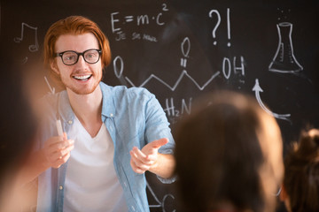 Young cheerful teacher in eyeglasses pointing at one of students giving right answer to his question at lesson