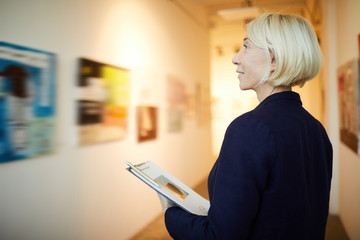 Side view portrait of mature female manager holding clipboard working in art gallery, copy space © Seventyfour