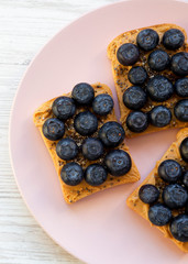 Vegan toasts with peanut butter, blueberries and chia seeds on a pink plate over white wooden background. Flat lay, from above. Close-up.