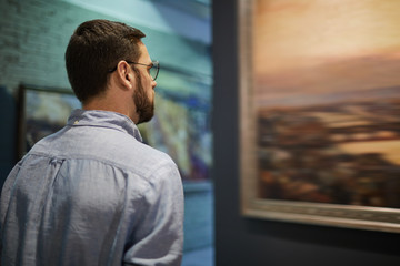 Back view portrait of young bearded man looking at pictures in modern art gallery or museum, copy...