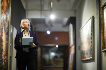 Portrait of mature woman holding clipboard looking at paintings standing in art gallery or museum,...