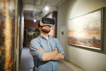 Waist up portrait of modern man wearing VR headset during virtual tour in art gallery or museum,...
