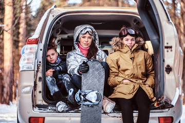 Happy family on vacation during the winter holidays. A beautiful woman and her sons dressed in warm clothes sitting on the trunk of a car