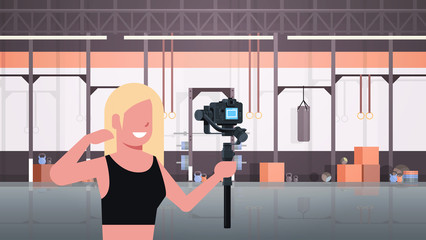 woman athlete fitness blogger shooting selfie video girl in front of DSRL camera recording herself using motorized gimbal stabilizer social media concept modern gym interior horizontal