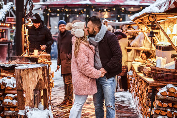 A happy couple in love wearing stylish warm clothes holding hands and kissing while standing at the winter fair at a Christmas time.