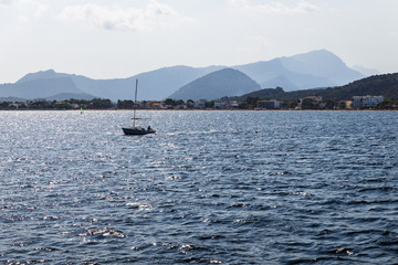 wide, blue sea waters on a sunny day, mountainous and shoreline in the distance; a lonely boat with a lowered sail