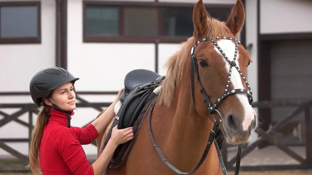 Pretty girl preparing saddle for riding horse. Slow motion young attractive woman in helmet preparing her horse for riding at ranch.