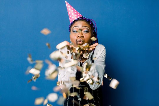 beautiful young black woman celebrating birthday or new year and chrismas party while blowing confetti decorations to camera isolated over blue background