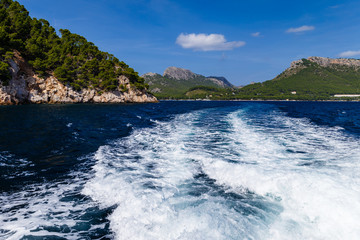 Obraz premium sunny day, sea view to the coast; view of the white waves left by the motorboat; on the shore there are large mountains partially overgrown with trees