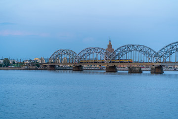 Naklejka premium Widok na panoramę miasta z mostem kolejowym i pociągiem na nim w Rydze, Łotwa, w Blue Hour, Zmierzch z wolną przestrzenią na tekst, Koncepcja podróży