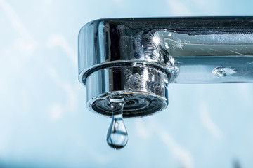 A drop of water hangs out of a modern chrome plated water tap faucet macro close-up.