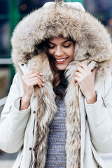 Attractive stylish girl dressed in a knitted dress and a light coat with fur poses in the street