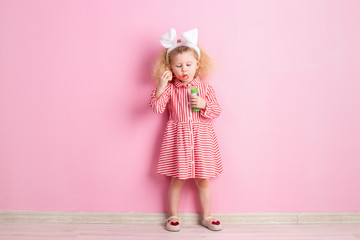 Lovely little girl in a striped red and white dress and bunny ears on her head inflates soap bubbles standing against a pink wall