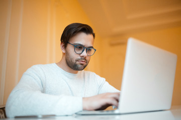 Serious young man working on laptop over yellow background. Dressed in white sweater and wearing blue glasses.