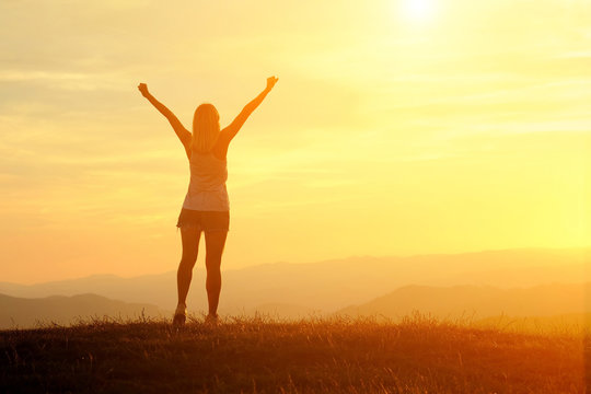 Happy woman traveler with open arms stay on the peak of mountain in sunset