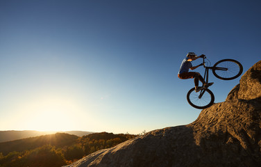 Fototapeta na wymiar Cyclist standing on back wheel on trial bike. Professional sportsman rider making acrobatic stunt on the edge of big boulder on the top of mountain at sunset. Concept of extreme sport active lifestyle