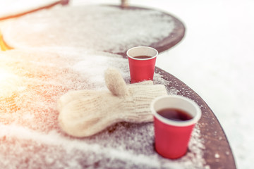 Obraz na płótnie Canvas Wooden table covered with snow in winter in the city. Two cups of coffee and tea. Warm mittens on the rack. Free space for text. The concept of breakfast or lunch on the street.
