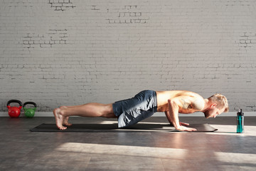 Muscular athlete exercising yoga asana plank in sunny sport club, brick wall. Fit shirtless male...