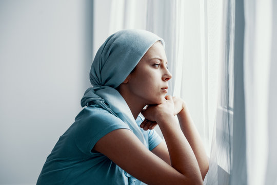 Thoughtful young girl suffering from bone cancer, wearing blue headscarf and looking through the window in hospital after surgery