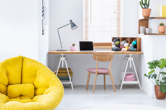 Yellow pouf in white home office interior with wooden chair at desk with lamp and laptop. Real photo