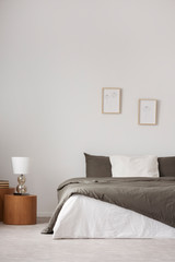 Lamp on wooden stool next to bed in white minimal bedroom interior with posters. Real photo
