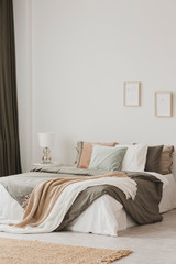 Blanket and pillows on bed in white minimal bedroom interior with rug and posters. Real photo