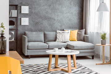 Round coffee table on patterned rug in tasteful living room with grey sofa with pillows, copy space on empty concrete wall