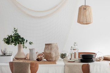 Green plant in beige vase, white jug and wicker lantern on dining table in bright interior - obrazy, fototapety, plakaty