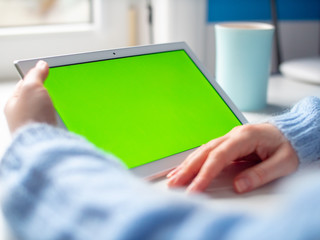 Woman hand hold white tablet with green screen and coffee cup in office on white table. Close up.